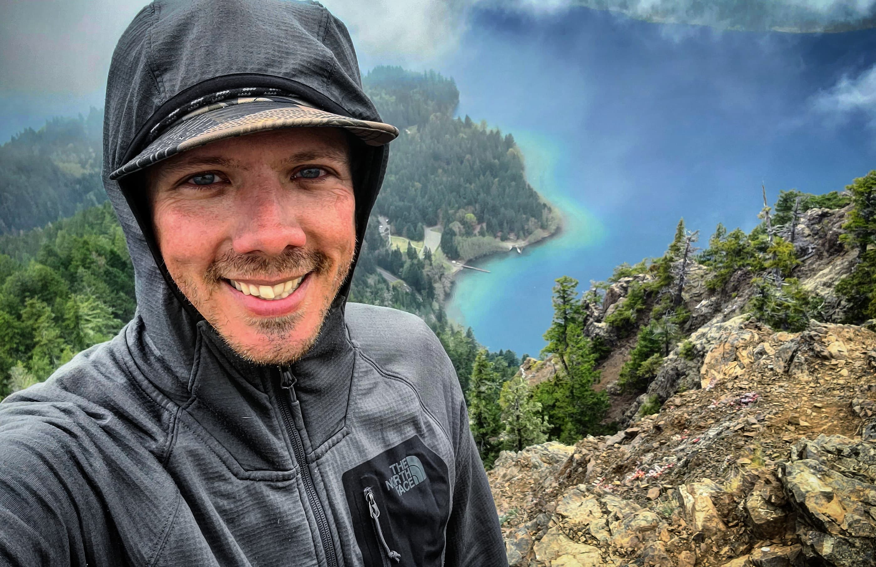 Smiling man in a grey hoodie overlooking a scenic blue lake from a rocky cliffside.